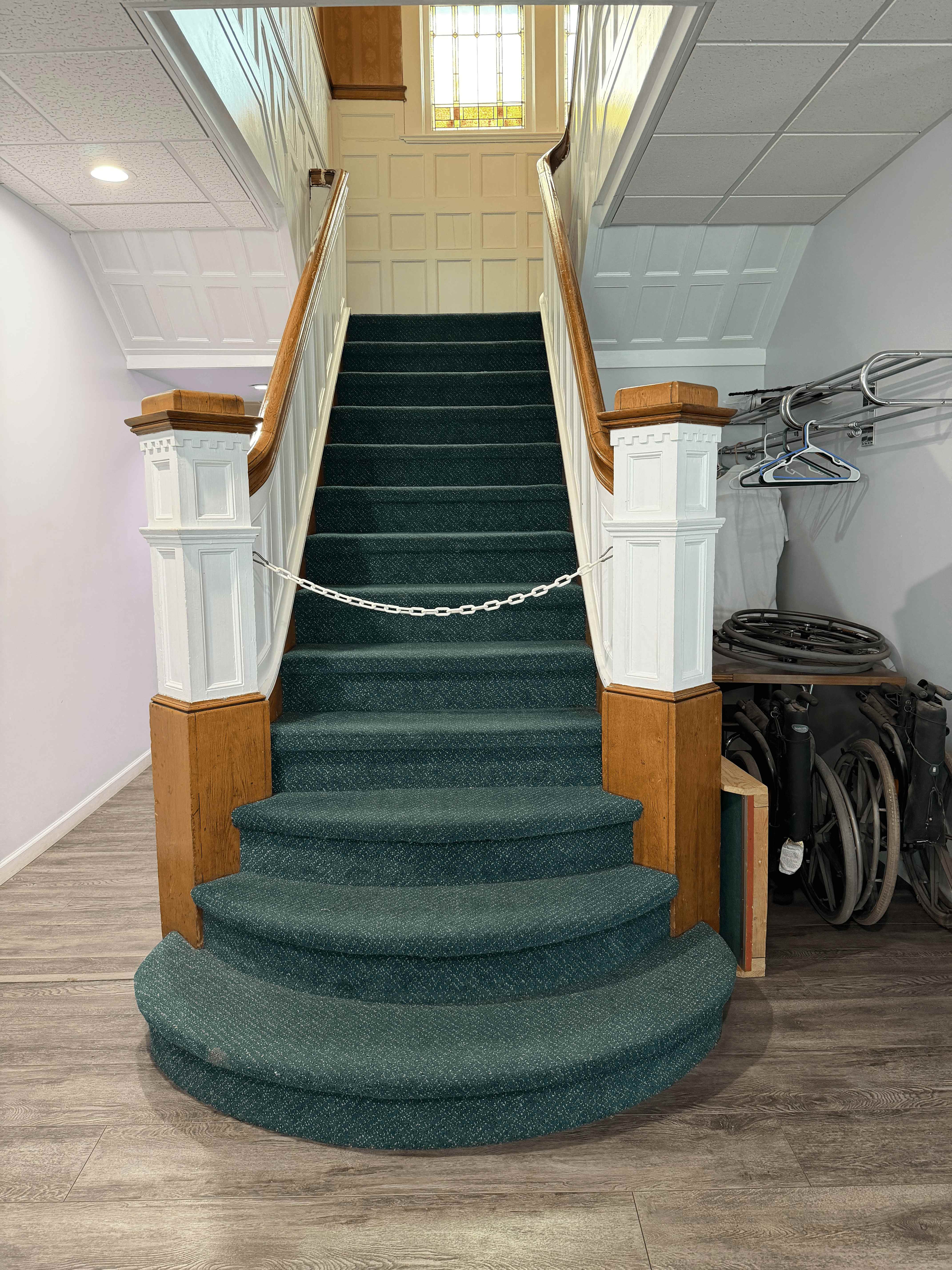 Green-carpeted grand staircase with wood newel posts and a stained-glass window above