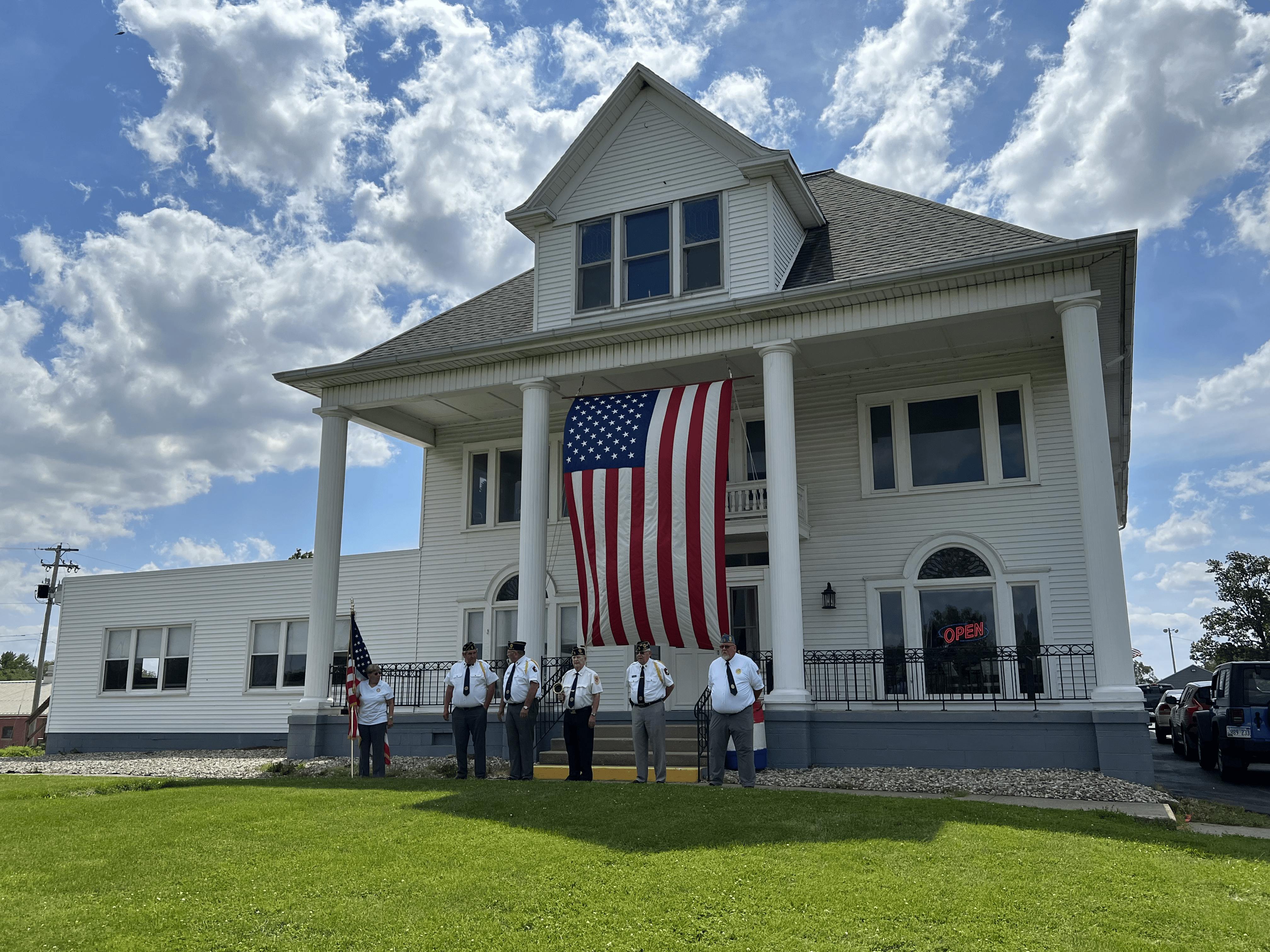 American Legion Post #211 members in uniform standing in front of the Post building with a large American flag draped from the porch